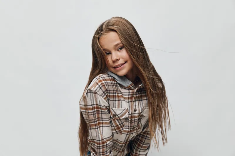 Girl with long hair in plaid shirt smiling and posing against plain background. Child with casual clothes and confident expression in studio photography session.