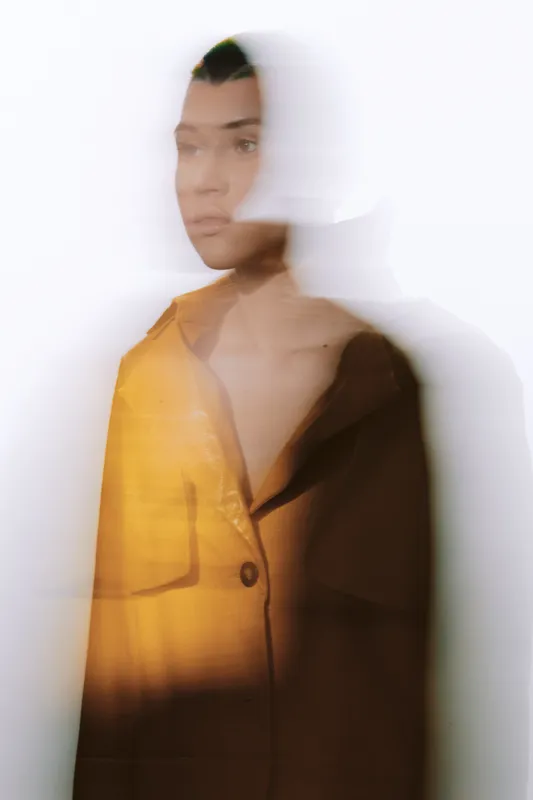 Young woman with motion blur effect in yellow and dark jacket, creative portrait with soft lighting and abstract white background, artistic and modern photography style.