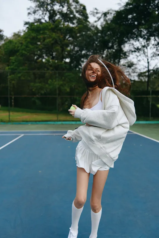 Joyful female athlete in a white sports outfit and hoodie, celebrating on a tennis court surrounded by lush greenery, embodying a vibrant and energetic spirit