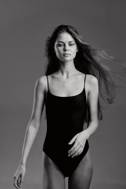 Black and white image of a young woman in a black bodysuit with flowing hair and a calm expression.