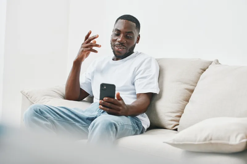 Happy young Black man relaxing on a couch while using a smartphone, dressed in a casual white t shirt and jeans, exuding a positive vibe in a minimalist interior