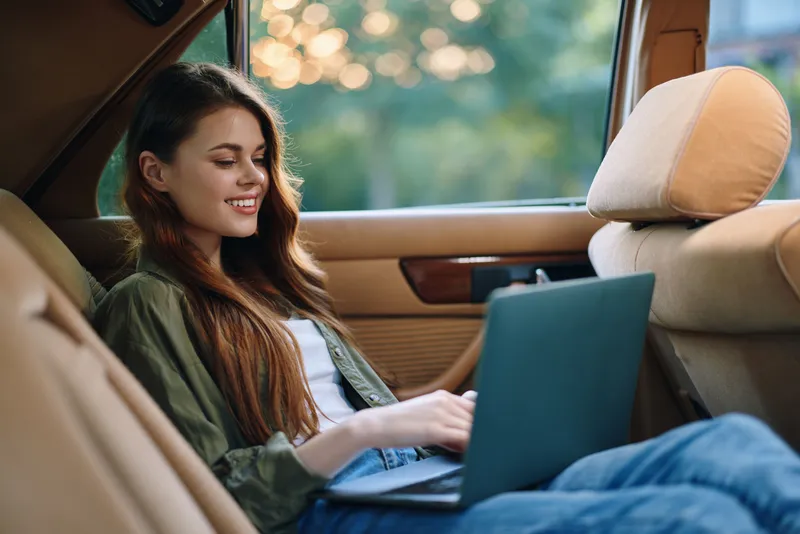Young woman enjoying remote work in a cozy car interior, smiling while using a laptop, showcasing a relaxed and productive vibe, perfect for lifestyle and freelance themes.