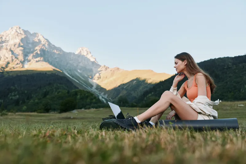 A focused young woman sits outdoors in a grassy meadow with a laptop, brainstorming ideas. She rests her chin on her hand, smiling slightly, while the tranquil mountains create a serene, productive