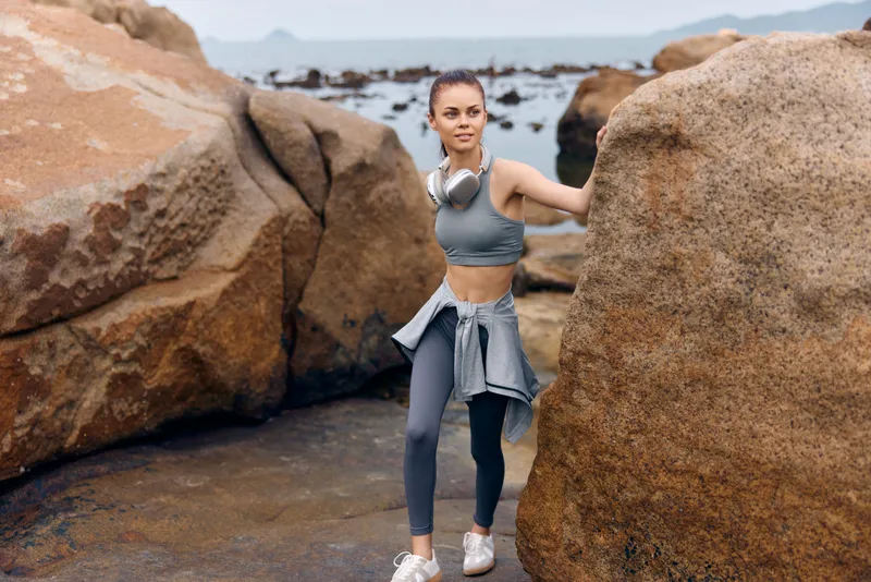 Athletic young woman in stylish activewear navigating rocky coastline, showcasing a vibrant sea background and a healthy lifestyle