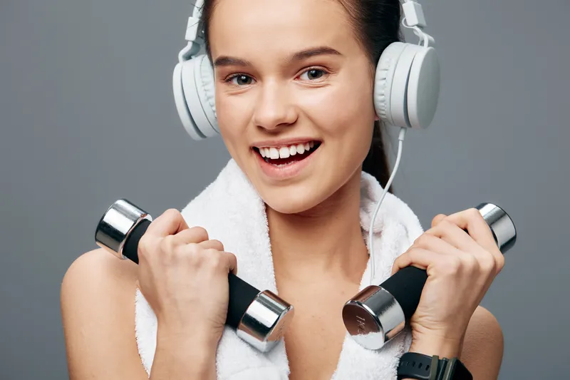 Woman in fitness theme lifting dumbbells, wearing headphones and a towel around neck, smiling, focused on strength and music in a gym setting, energetic workout moment.
