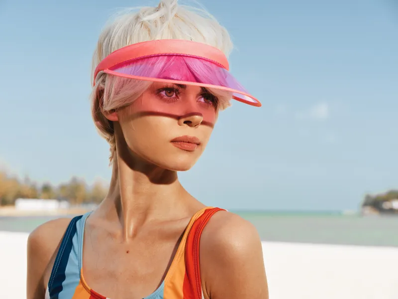 Bright beach portrait of a young woman wearing a pink translucent visor, sunny skies, turquoise water, sunlit skin, and a stylish sporty vibe.