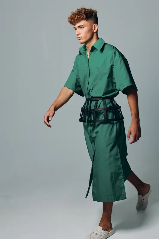 Young man walking in studio wearing green oversized shirt and matching skirt with black belt, styled with white slip-on shoes on plain background.