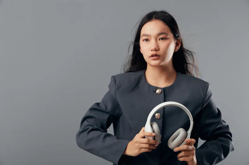 Young woman in formal blazer holding wireless headphones with neutral expression on gray background studio. Portrait of Asian female with long hair and casual technology accessory.