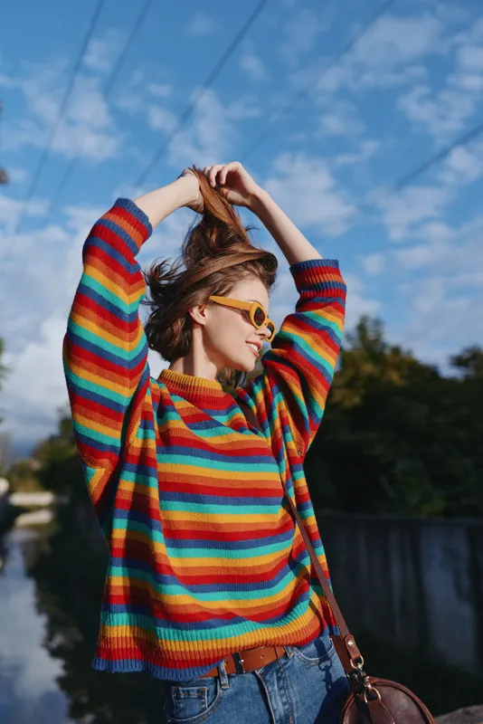 Woman striped sweater sunglasses outdoor jeans casual smiling urban young woman in her 20s pulling hair up, rainbow knit, yellow shades, brown shoulder bag, sunny lifestyle travel portrait.