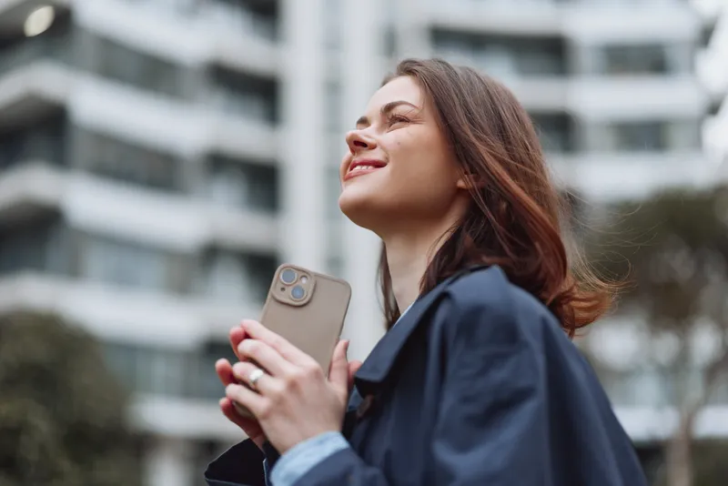 Cheerful woman outdoors, smiling with eyes closed, sunlit moment, relaxed posture, soft breeze, natural colors, urban background, modern architecture