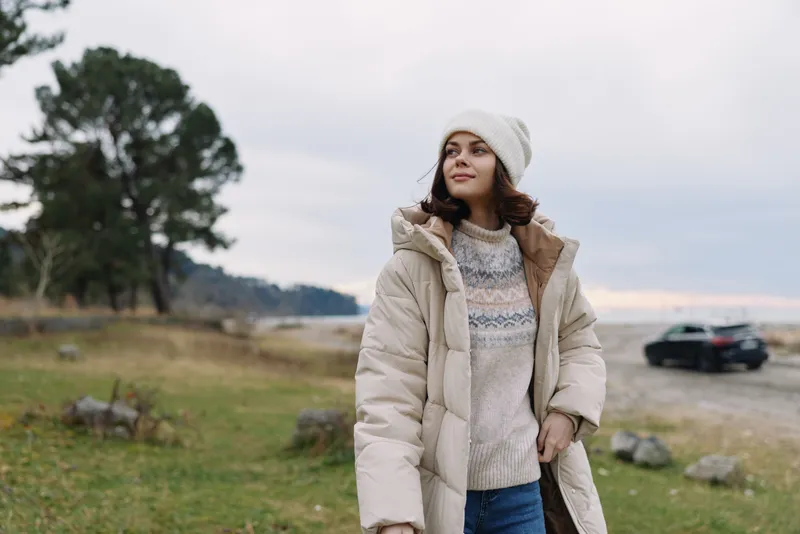 Winter outdoors, a woman wearing a cozy coat and knit sweater beanie strolling in an open landscape with trees and muted sky conveying calm seasonal fashion and natural serenity