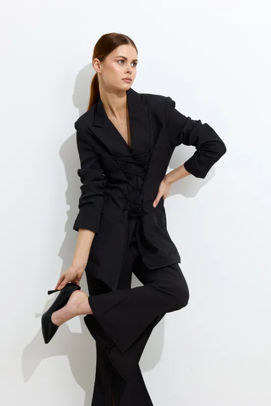 Fashion woman wearing a stylish black blazer with lace-up detail and matching pants, posing confidently against a white background in modern minimalistic style.