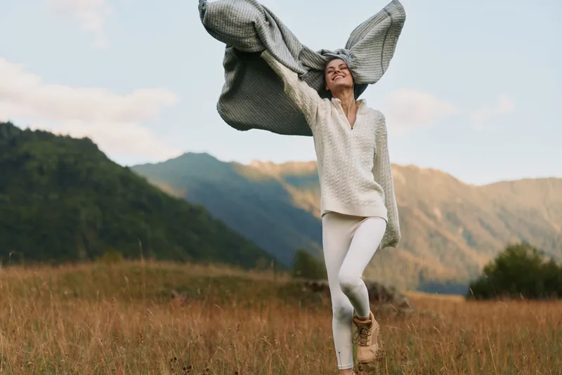 Woman dances in a field near mountains, wearing white sweater and pants, scarf billowing, carefree lifestyle shot capturing joy, movement and outdoor freedom