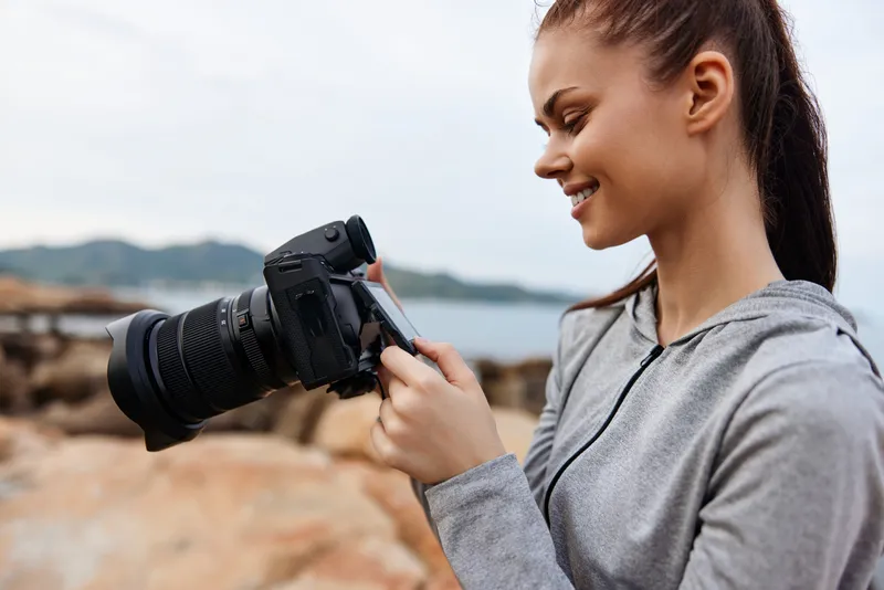Young woman capturing nature photos while enjoying photography on the rocky beach, wearing a casual grey hoodie, smiling in a coastal setting