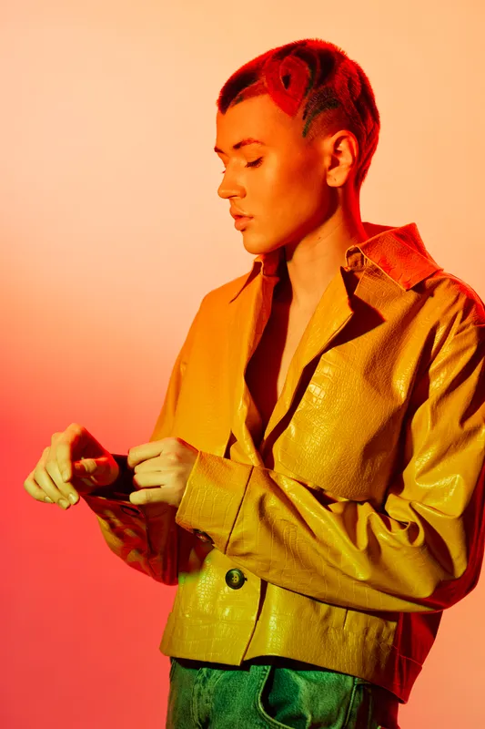 Young woman with red hair wearing a yellow jacket poses thoughtfully against an orange-red background, highlighting fashion, style, and modern expression.