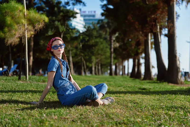 Lifestyle woman in modern boho-western denim street style sitting on grass outdoors wearing sunglasses and a headband, enjoying sunny weather with film color tones.