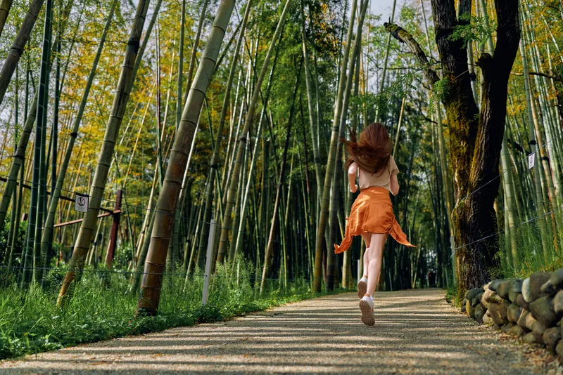 Woman running barefoot on gravel path through bamboo forest, back view of girl in orange dress jogging along tree-lined trail, nature motion and outdoor freedom in sunlight.