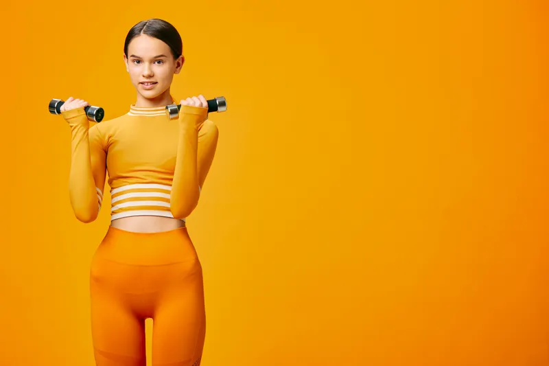 Fitness woman in an orange athletic set lifts dumbbells against a bold orange backdrop, showcasing strength, focus, and healthy living in a modern workout scene.