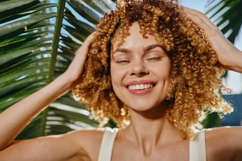 Smiling woman with curly hair enjoys sunlight outdoors near green palm leaves, expressing happiness and natural beauty in warm daylight.