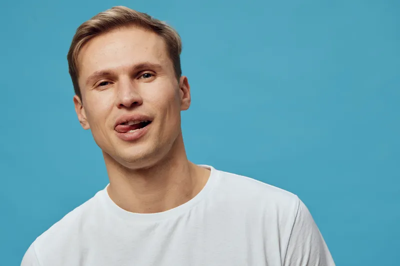 Close-up of a funny young man with light skin, short brown hair, wearing casual white t-shirt, making a silly face, isolated on plain blue background. People lifestyle concept, entertainment, humor, f