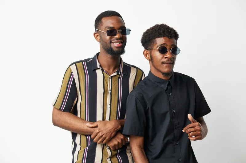 Happy African American men wearing sunglasses pose together confidently, showcasing stylish outfits and a playful vibe. Their friendship radiates positivity and style.