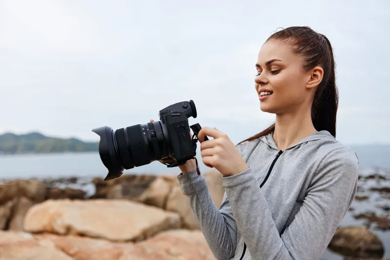 Young woman with a camera by the sea, capturing moments in a gray hoodie, expressing joy and creativity amidst a natural coastal background