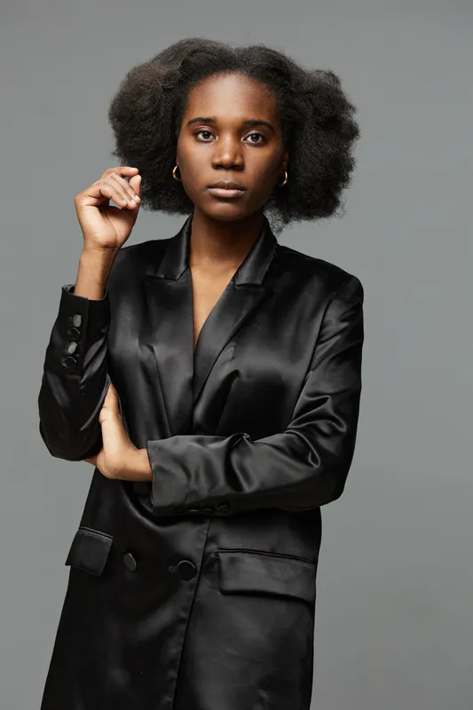 Portrait of a confident Black woman in a satin black blazer, with natural curly hair, showcasing a serious expression against a neutral gray background