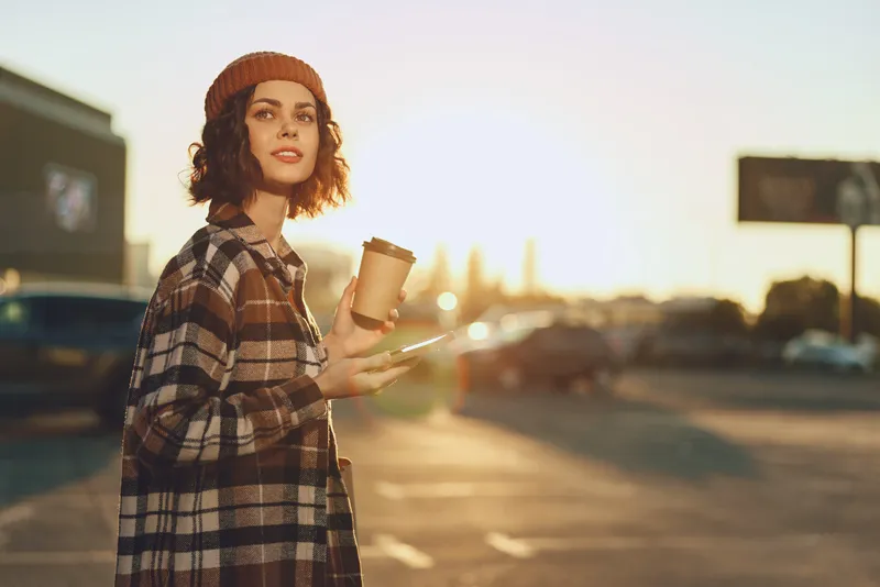 Woman with coffee and phone in urban street at golden hour, candid smile and beanie conveying authenticity, mindful living and emotional storytelling in a modern city lifestyle.