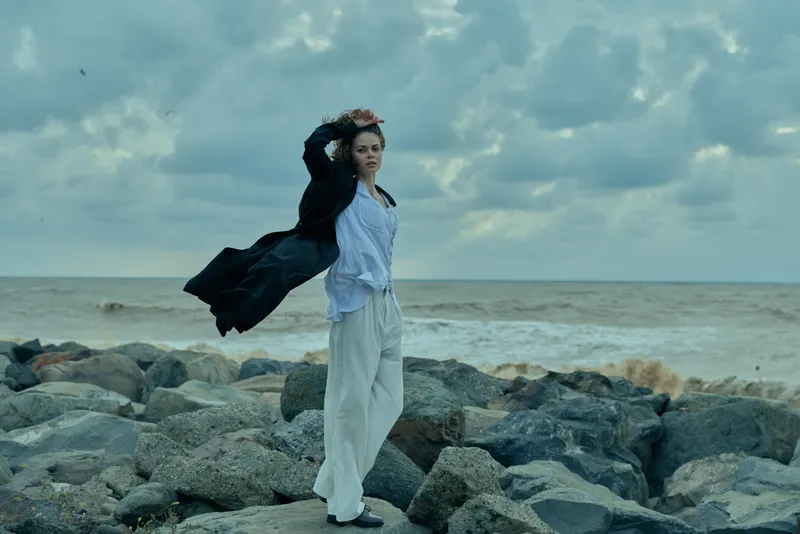 Woman standing on a rocky shoreline with flowing coat, windy sea backdrop, casual fashion pose, dynamic movement, overcast sky, ocean waves, outdoor scene, moody lifestyle photography