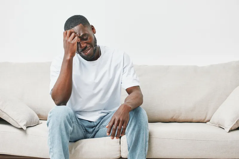 A young African American man sitting on a light sofa, showing a range of emotions, dressed casually in a white t shirt and denim jeans