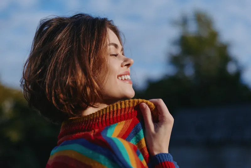Young woman in her 20s smiling outdoors, wearing a rainbow sweater, eyes closed and holding the collar, joyful expression in sunlight for lifestyle and travel imagery