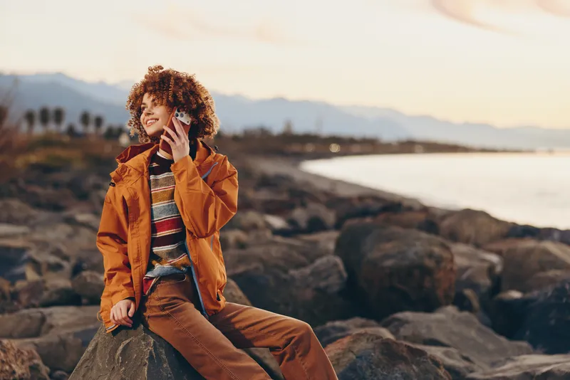 woman lifestyle seaside rocks sunset jacket curly hair smile coast, woman sits on rocky shore near calm sea at sunset, casual jacket and striped sweater, natural smile, peaceful travel moment