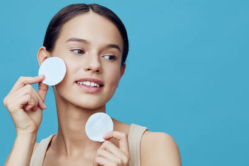 Young woman with clear skin applying cotton pads for skincare against a blue background, portraying beauty and freshness, ideal for health and beauty concepts