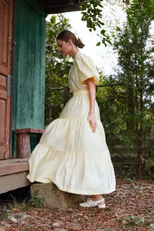 Woman in a pale yellow dress walks outdoors beside a wooden doorway, capturing graceful movement and a serene garden atmosphere with soft natural light.