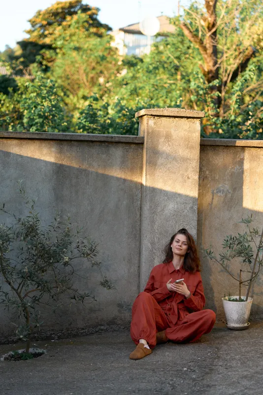 Woman seated in a red dress in an outdoor garden, showcasing relaxation and lifestyle. Soft daylight highlights the garden backdrop and stone wall