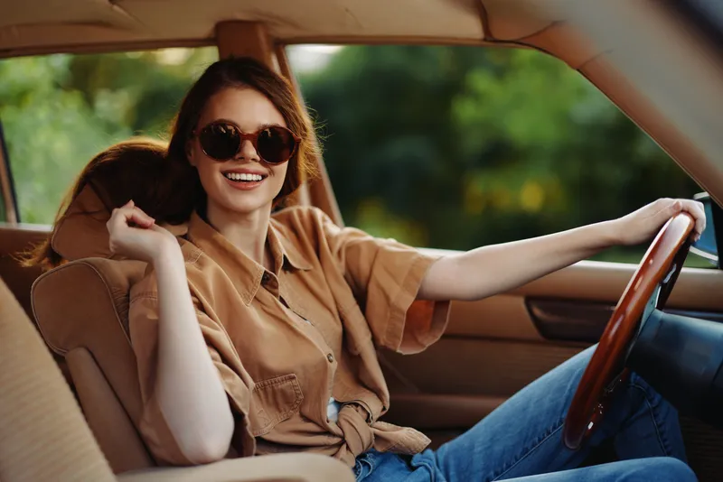 Young woman smiling in a vintage car, wearing sunglasses and a casual shirt, capturing a joyful summer vibe with greenery in the background.