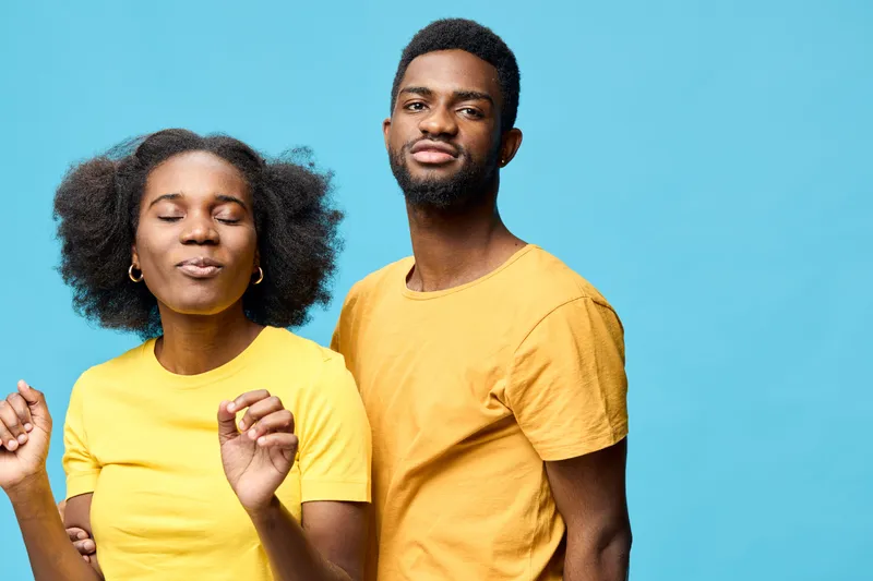 Joyful young African couple dancing together in matching yellow t shirts against a vibrant blue background, radiating happiness and fun during a warm season