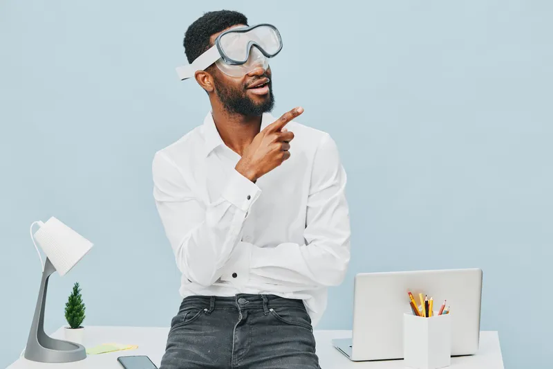 Person wearing safety goggles and pointing upward, working in modern office with laptop, stationery, and desk accessories, blue wall background. Creative workplace concept.