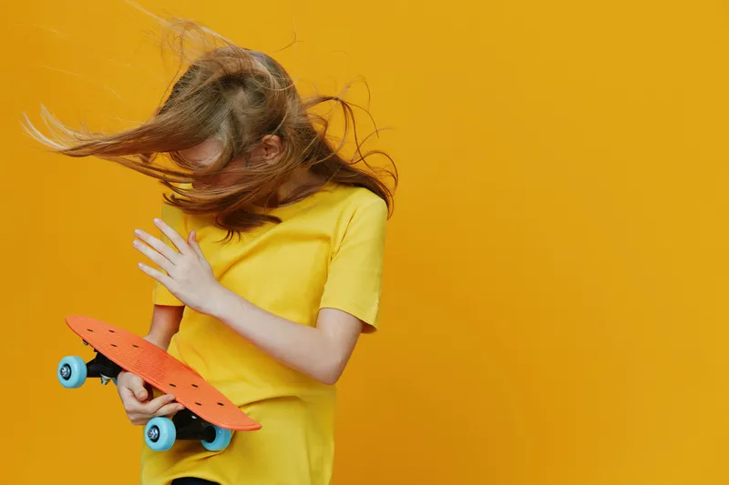 Youthful girl with long hair in a yellow shirt holding a skateboard, expressing joy against a vibrant yellow background, ideal for sports and lifestyle themes