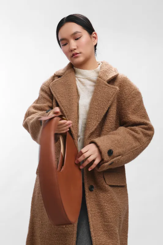 asian woman in a warm tailored coat adjusts a leather bag, standing in a clean studio with soft lighting, creating a calm, elegant fashion moment that feels polished and contemporary.