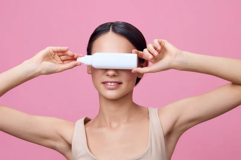 A young woman with dark hair holds a white bottle in front of her eyes, representing beauty and skincare concepts against a pink background