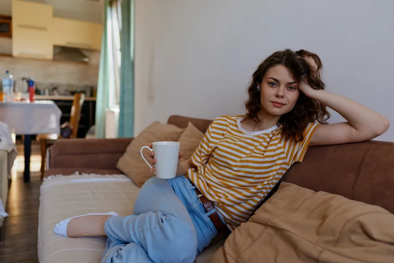 A young woman relaxes on a sofa at home, holding a mug and gazing thoughtfully into the distance, capturing a casual, reflective moment in a cozy living space.
