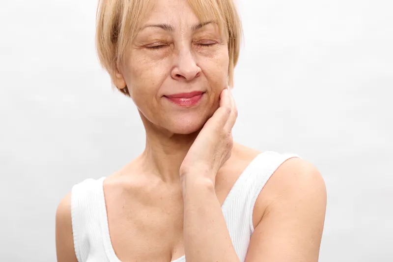 Senior woman with closed eyes touching chin, pained expression, soft white studio background, concept of facial pain and stress, emotional strain from aging