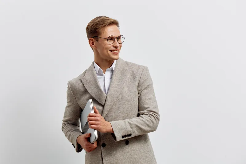 Confident woman dressed in a beige blazer smiling, holding notebook and pen, looking away, standing against plain gray background in studio. Business, professional, education, success, portrait, femal