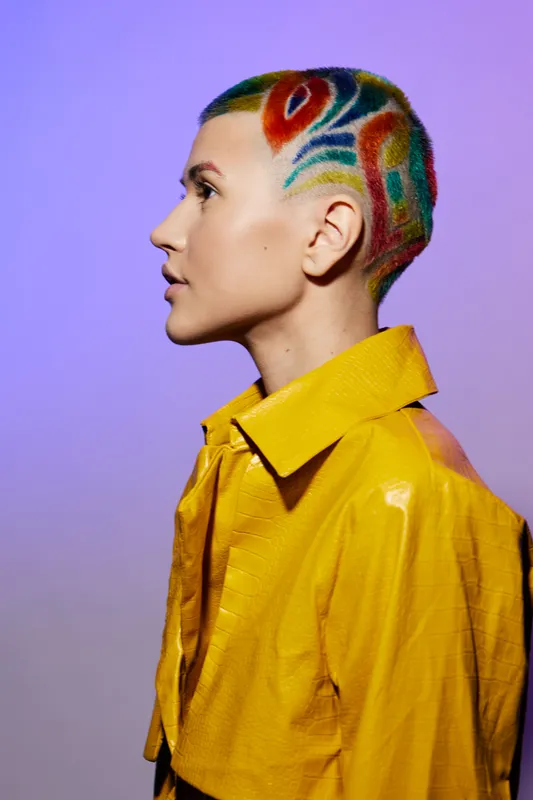 Woman with colorful shaved hairstyle wearing a vibrant yellow jacket posing against purple background, showcasing bold fashion and modern artistic expression.