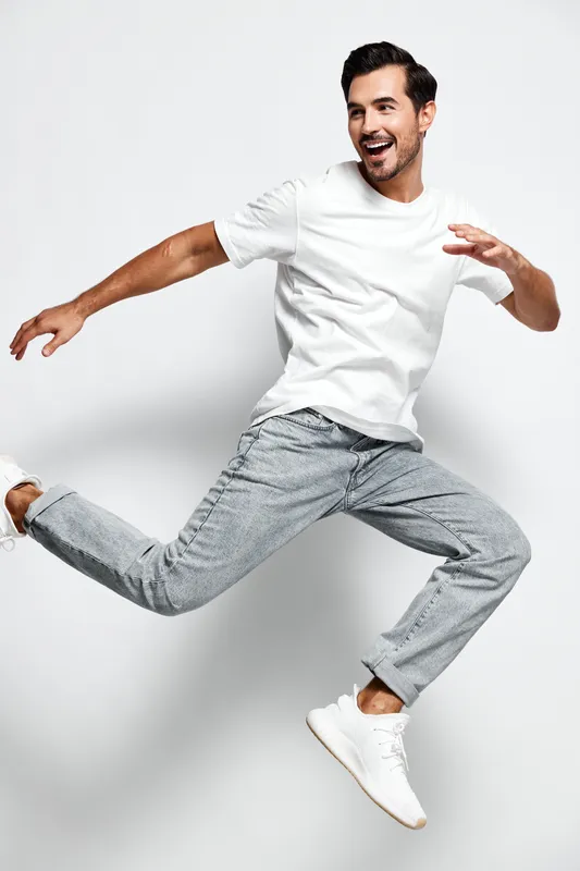 Full body smiling happy cheerful young man wearing white t-shirt jumping in the air, showcasing dynamic movement and joy against a plain gray background.
