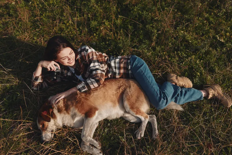 Woman lying on grass with a dog beside her, holding a camera in a relaxed outdoor scene, natural light, calm countryside vibe, candid moment of companionship.