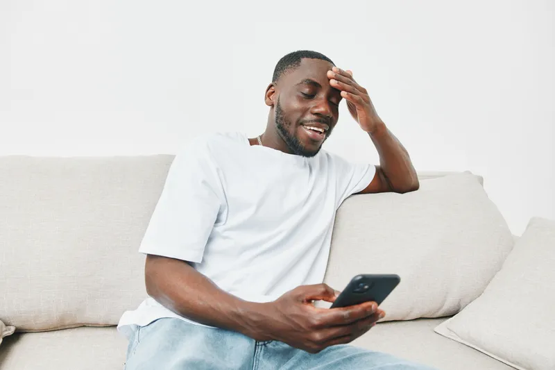 Happy young man relaxing on a sofa while checking his phone, wearing a casual white t shirt, showcasing a modern and comfortable lifestyle