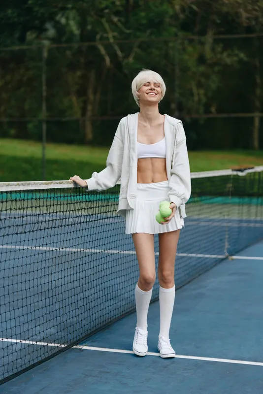 Young female tennis player smiling on the court, wearing a sporty white outfit, holding a green tennis ball, surrounded by a natural green background