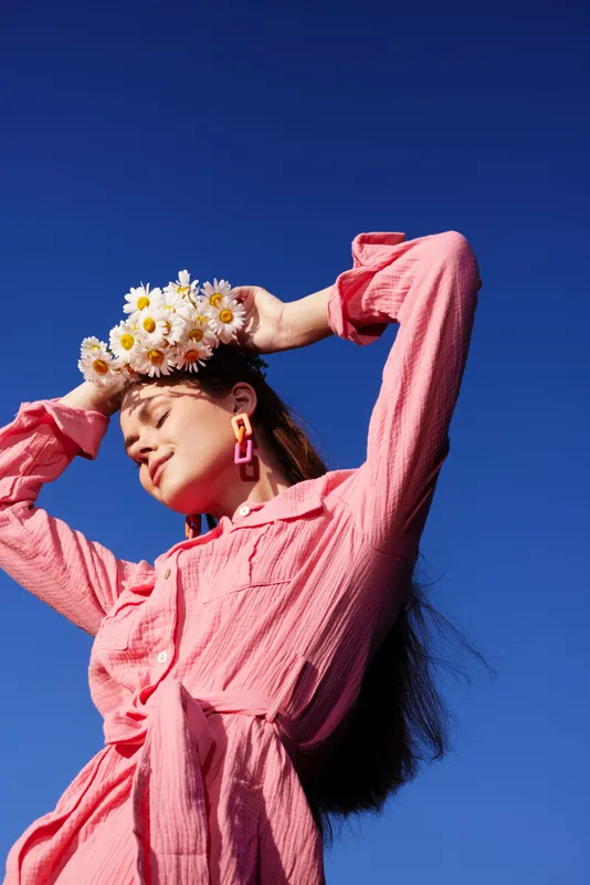 Young woman with flower crown enjoying the sun in a pink dress against a clear blue sky, expressing joy and tranquility with vibrant colors